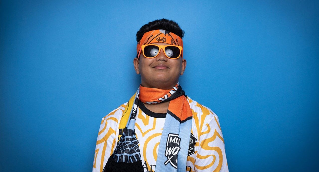 Arav Patil穿戴休士顿DynapFC设备,包括橙色太阳镜、头带、工具箱和围巾,同时站立蓝底