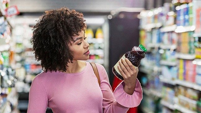 卷黄头发和粉红色衬衣的女人站在杂货店走道上看一瓶紫果汁的营养信息